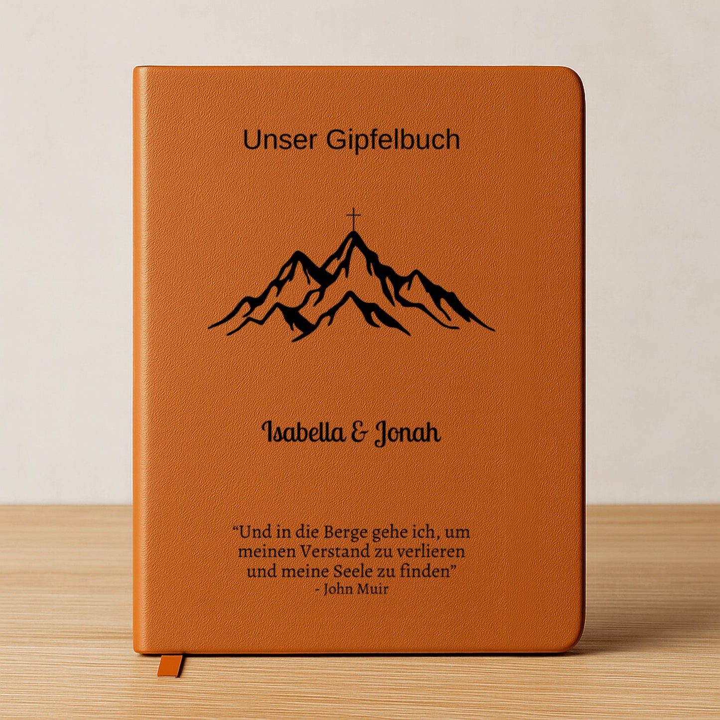 Ein personalisiertes Gipfelbuch in A5 mit einem braunen, veganen Ledereinband liegt auf einer hellen Oberfläche. Auf dem Cover sind in Schwarz ein Bergmotiv mit einem Gipfelkreuz und der Schriftzug "Unser Gipfelbuch" zu sehen. Darunter stehen die personalisierten Namen "Isabella & Jonah" sowie ein inspirierendes Zitat von John Muir über die Berge.