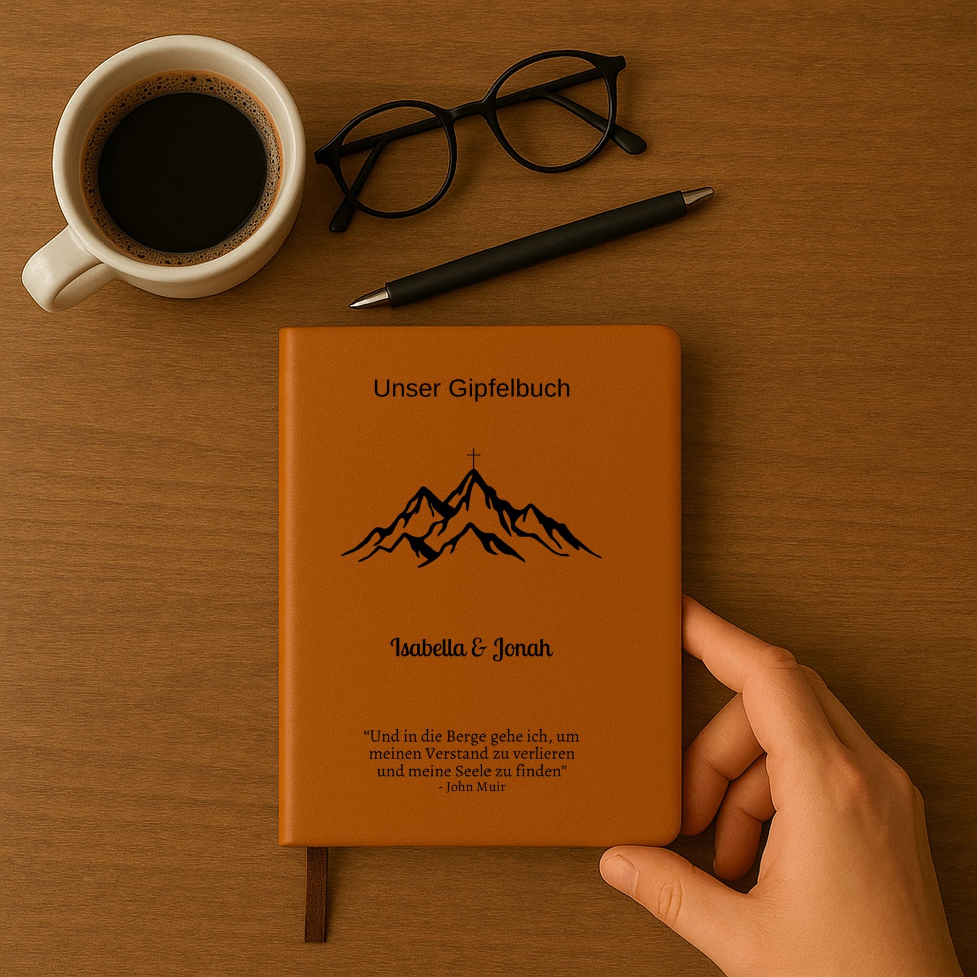 Ein personalisiertes Gipfelbuch in A5 mit einem braunen, veganen Ledereinband liegt auf einer hellen Oberfläche. Auf dem Cover sind in Schwarz ein Bergmotiv mit einem Gipfelkreuz und der Schriftzug "Unser Gipfelbuch" zu sehen. Darunter stehen die personalisierten Namen "Isabella & Jonah" sowie ein inspirierendes Zitat von John Muir über die Berge.