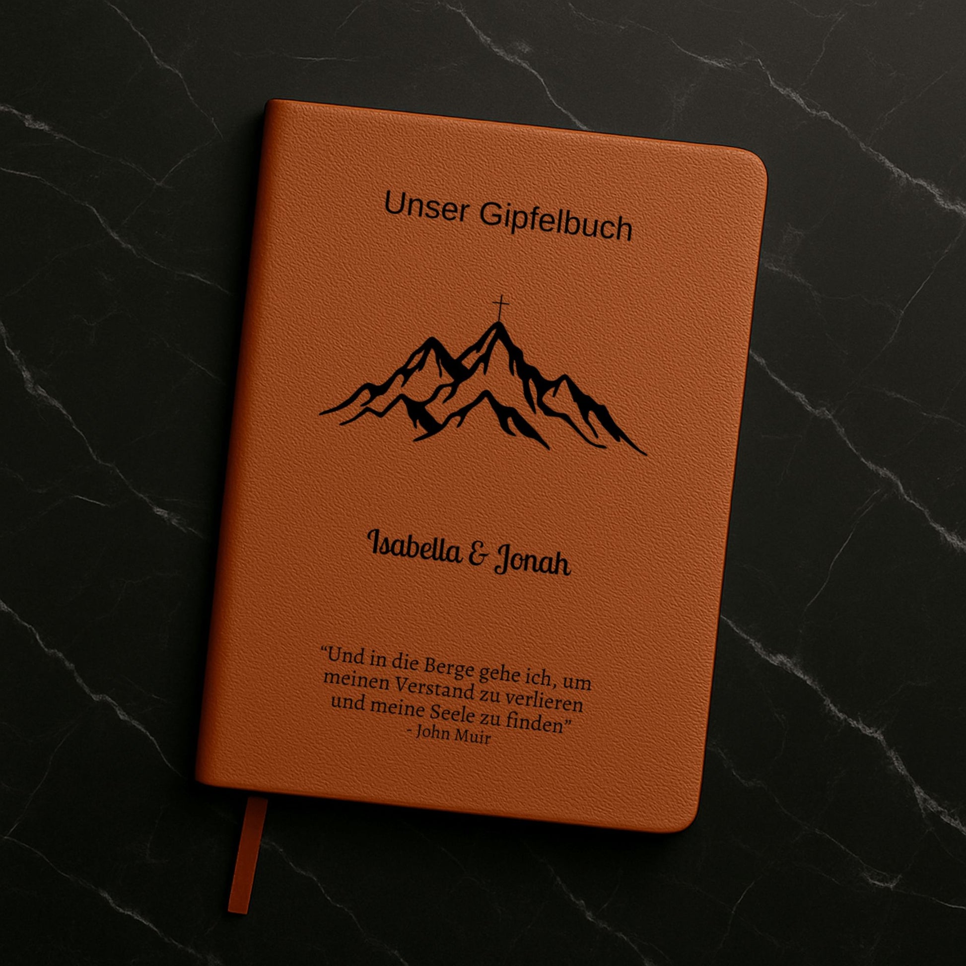 Ein personalisiertes Gipfelbuch in A5 mit einem braunen, veganen Ledereinband liegt auf einer hellen Oberfläche. Auf dem Cover sind in Schwarz ein Bergmotiv mit einem Gipfelkreuz und der Schriftzug "Unser Gipfelbuch" zu sehen. Darunter stehen die personalisierten Namen "Isabella & Jonah" sowie ein inspirierendes Zitat von John Muir über die Berge.