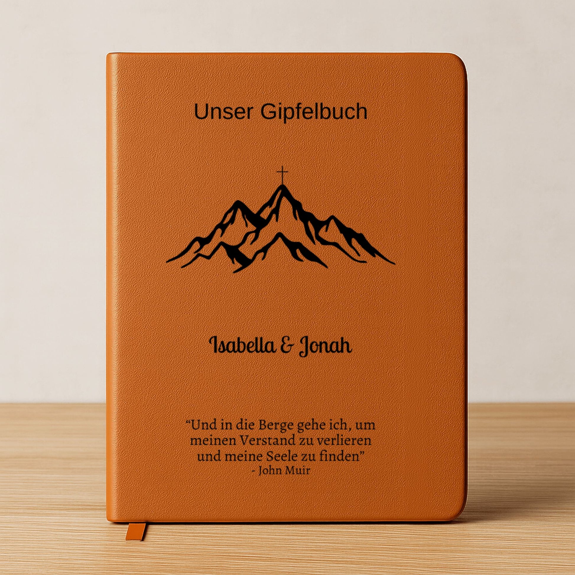 Ein personalisiertes Gipfelbuch in A5 mit einem braunen, veganen Ledereinband liegt auf einer hellen Oberfläche. Auf dem Cover sind in Schwarz ein Bergmotiv mit einem Gipfelkreuz und der Schriftzug "Unser Gipfelbuch" zu sehen. Darunter stehen die personalisierten Namen "Isabella & Jonah" sowie ein inspirierendes Zitat von John Muir über die Berge.