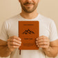 Ein personalisiertes Gipfelbuch in A5 mit einem braunen, veganen Ledereinband liegt auf einer hellen Oberfläche. Auf dem Cover sind in Schwarz ein Bergmotiv mit einem Gipfelkreuz und der Schriftzug "Unser Gipfelbuch" zu sehen. Darunter stehen die personalisierten Namen "Isabella & Jonah" sowie ein inspirierendes Zitat von John Muir über die Berge.