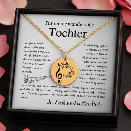 Eine goldene Halskette mit einem runden Anhänger, der mit einem Notenschlüssel und Musiknoten graviert ist. Die Kette wird in einer edlen, schwarzen Geschenkbox auf einer Karte mit einem inspirierenden, emotionalen Spruch präsentiert. Die liebevolle Aufmachung mit zarten Blütenblättern im Hintergrund positioniert den Schmuck als hochwertiges und bedeutungsvolles Geschenk für einen besonderen Menschen.