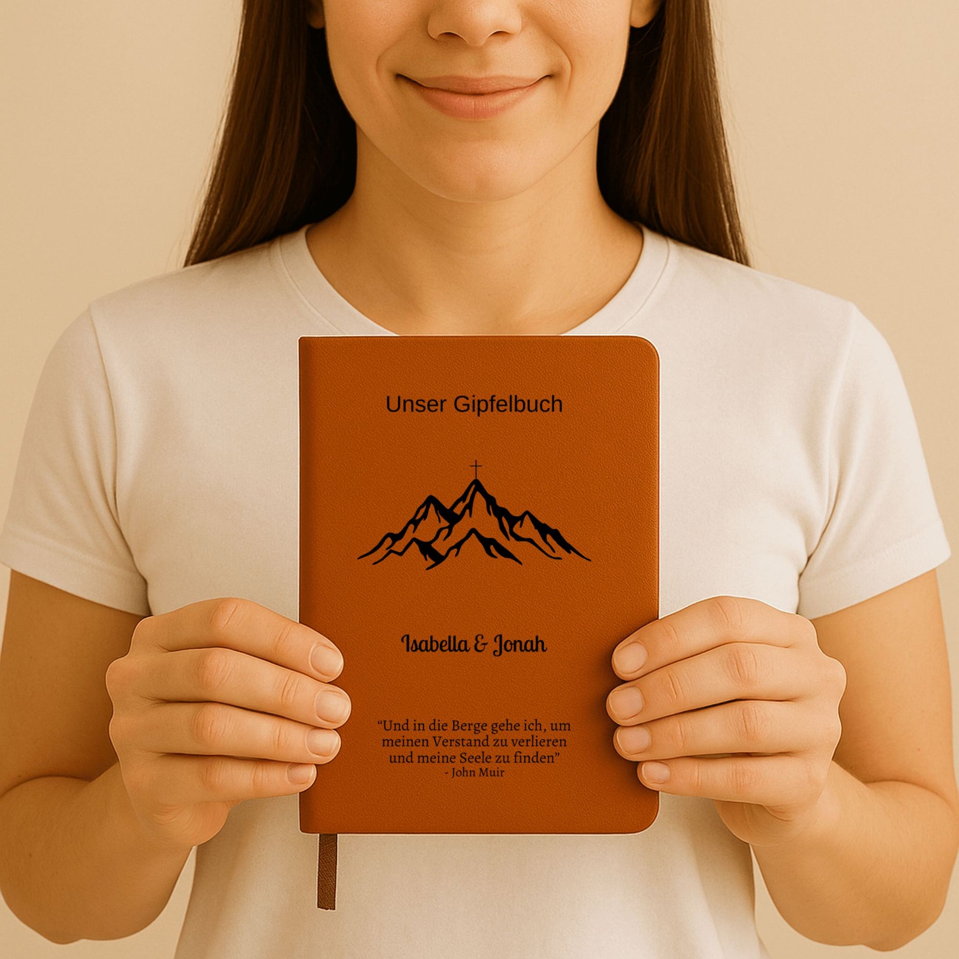 Ein personalisiertes Gipfelbuch in A5 mit einem braunen, veganen Ledereinband liegt auf einer hellen Oberfläche. Auf dem Cover sind in Schwarz ein Bergmotiv mit einem Gipfelkreuz und der Schriftzug "Unser Gipfelbuch" zu sehen. Darunter stehen die personalisierten Namen "Isabella & Jonah" sowie ein inspirierendes Zitat von John Muir über die Berge.