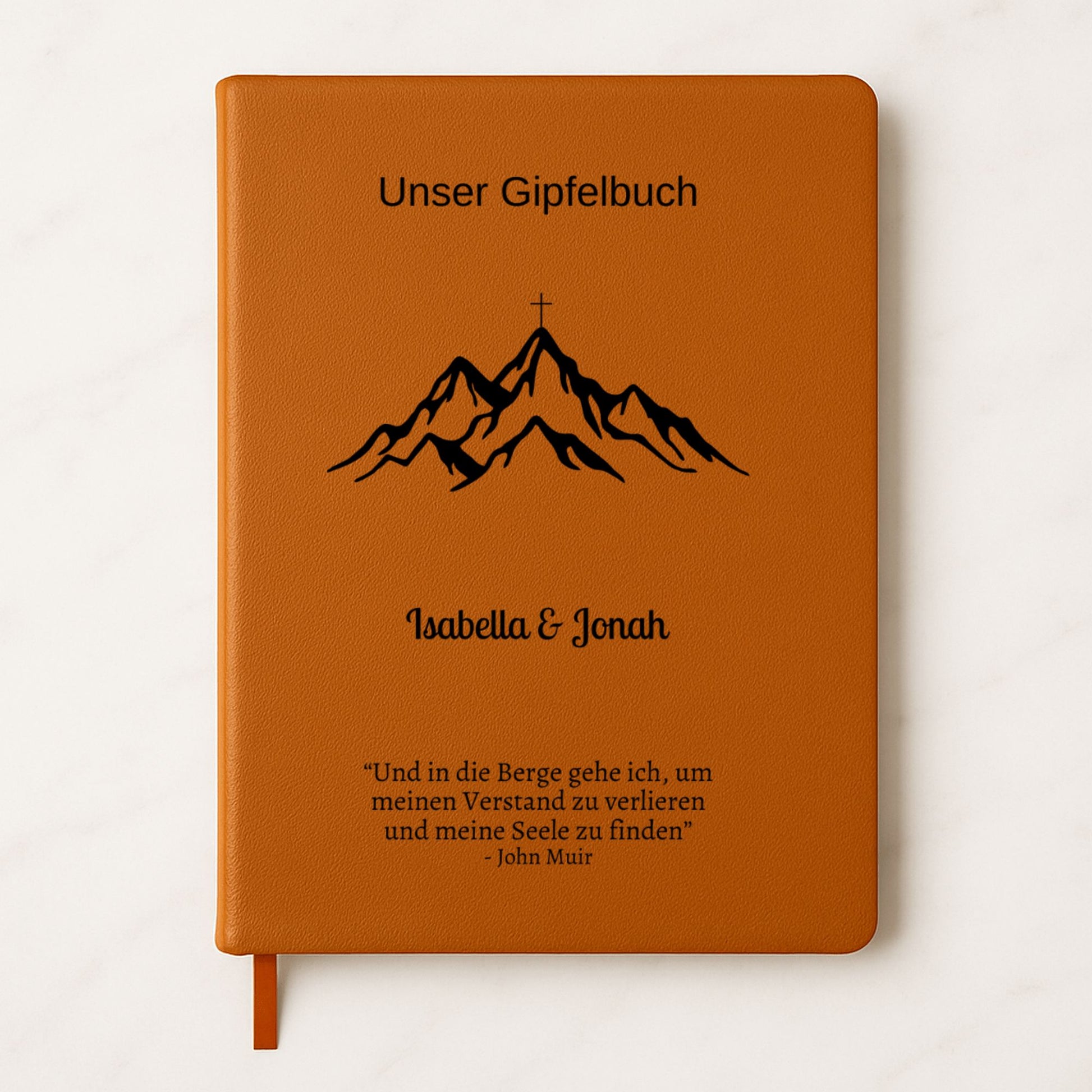 Ein personalisiertes Gipfelbuch in A5 mit einem braunen, veganen Ledereinband liegt auf einer hellen Oberfläche. Auf dem Cover sind in Schwarz ein Bergmotiv mit einem Gipfelkreuz und der Schriftzug "Unser Gipfelbuch" zu sehen. Darunter stehen die personalisierten Namen "Isabella & Jonah" sowie ein inspirierendes Zitat von John Muir über die Berge.