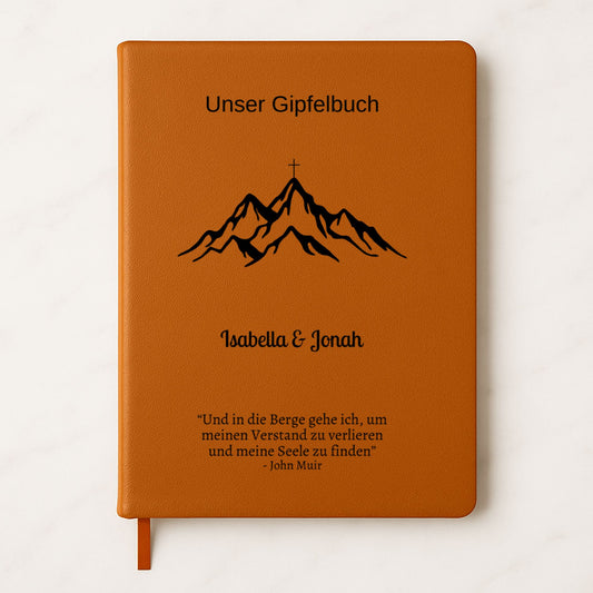 Ein personalisiertes Gipfelbuch in A5 mit einem braunen, veganen Ledereinband liegt auf einer hellen Oberfläche. Auf dem Cover sind in Schwarz ein Bergmotiv mit einem Gipfelkreuz und der Schriftzug "Unser Gipfelbuch" zu sehen. Darunter stehen die personalisierten Namen "Isabella & Jonah" sowie ein inspirierendes Zitat von John Muir über die Berge.