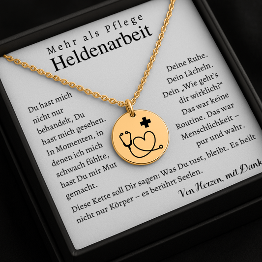 Goldene Halskette mit runden Anhänger, auf dem ein Stethoskop ein Herz bildet und ein kleines Plus-Zeichen daneben steht. Die Kette liegt in einer schwarzen Schmuckbox mit einer Dankeskarte. Auf der Karte steht: 'Mehr als Pflege – Heldenarbeit'. Umgeben von rosa Rosenblättern."