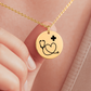 Goldene Halskette mit runden Anhänger, auf dem ein Stethoskop ein Herz bildet und ein kleines Plus-Zeichen daneben steht. Die Kette liegt in einer schwarzen Schmuckbox mit einer Dankeskarte. Auf der Karte steht: 'Mehr als Pflege – Heldenarbeit'. Umgeben von rosa Rosenblättern."