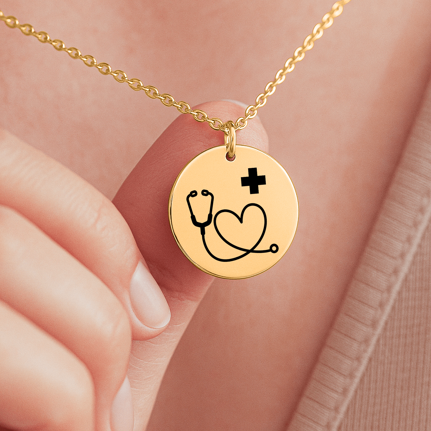 Goldene Halskette mit runden Anhänger, auf dem ein Stethoskop ein Herz bildet und ein kleines Plus-Zeichen daneben steht. Die Kette liegt in einer schwarzen Schmuckbox mit einer Dankeskarte. Auf der Karte steht: 'Mehr als Pflege – Heldenarbeit'. Umgeben von rosa Rosenblättern."